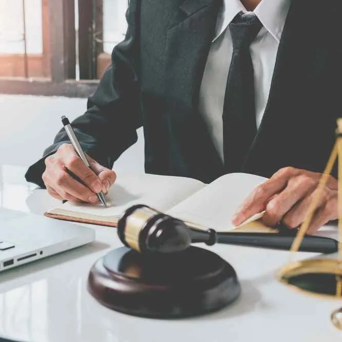Person in a black suit writing in a notebook with a judge's gavel and scales on the desk.