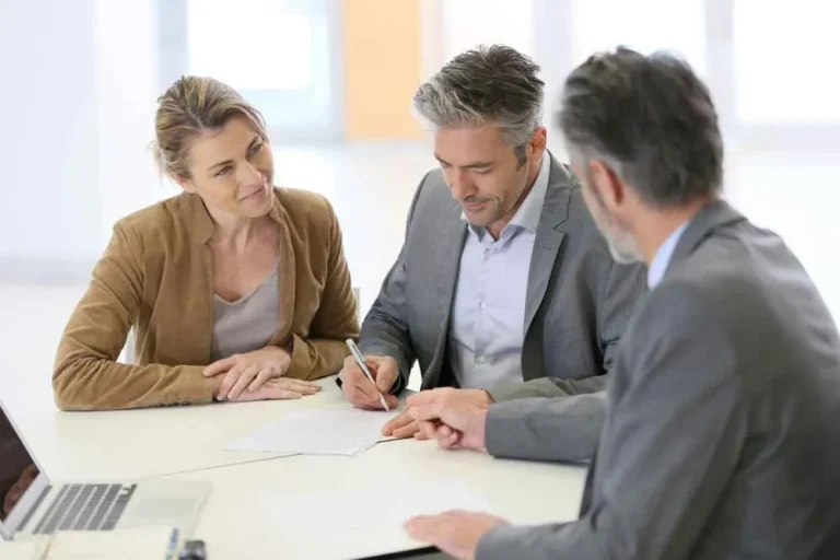 Three professionals in business attire seated at a white table, one man signing a document, another man and a woman observing.