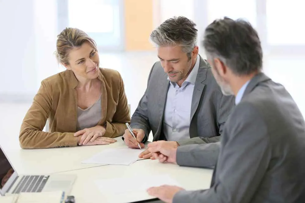 Three professionals in business attire seated at a white table, one man signing a document, another man and a woman observing.