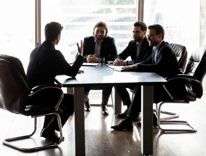 Four men in business attire engaged in a meeting around a black conference table in a bright office.