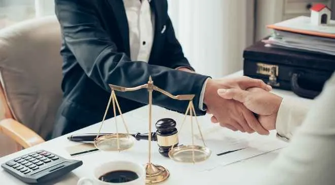 Two individuals in business attire shaking hands over a desk with legal scales, a gavel, documents, and a coffee cup.