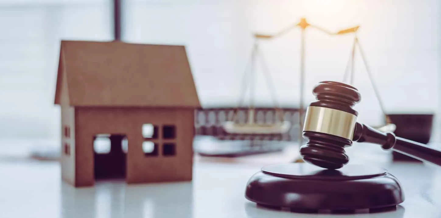 Wooden judge gavel beside miniature house model and blurred scales of justice.