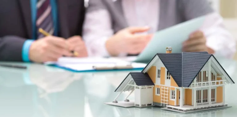 Scale model of a yellow house with black roof on glass table, two people reviewing documents.