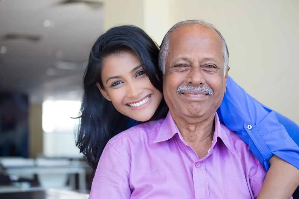 Smiling elderly man in pink shirt embraced from behind by younger woman in blue shirt.