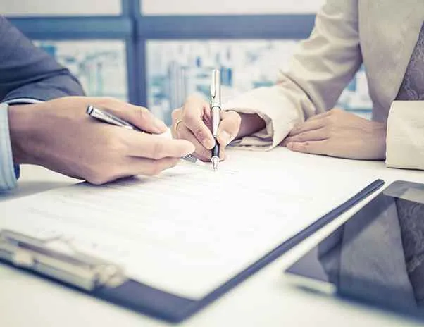 Two individuals in professional attire reviewing and signing documents on a clipboard at a desk.