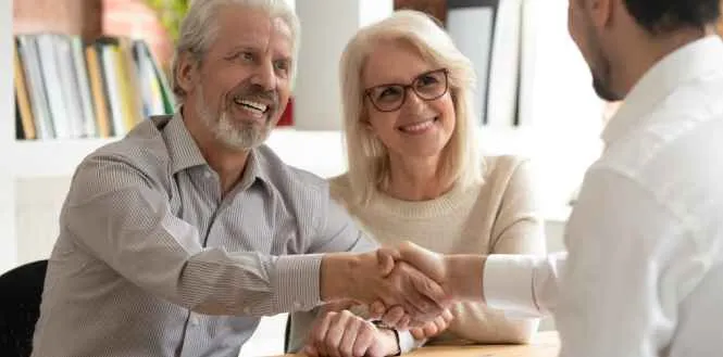 Two elderly individuals smiling and shaking hands with a professional in a bright office.