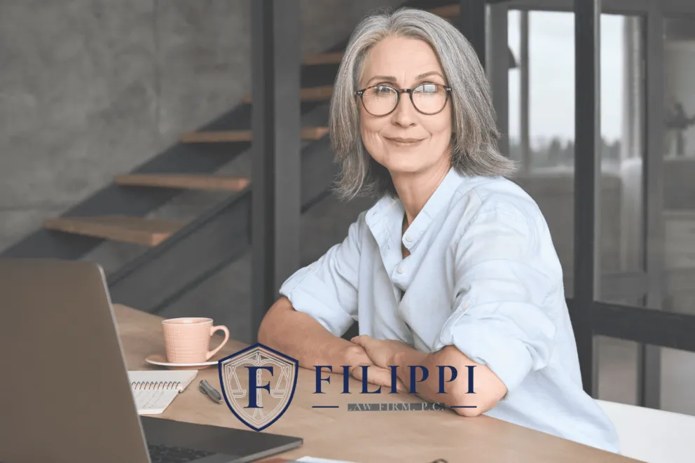 Professional middle-aged woman with gray hair and glasses seated at desk with laptop and coffee cup.