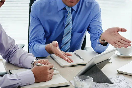 Two professionals in dress shirts and ties engaged in discussion over documents and a tablet at a meeting table.