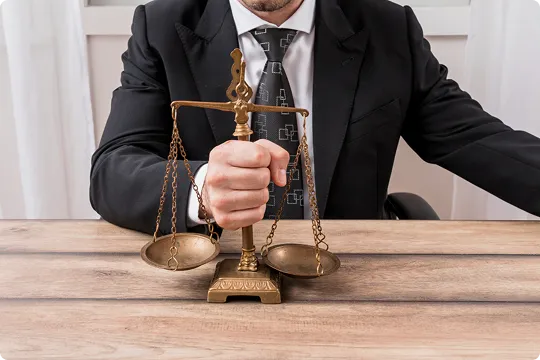 Man in black suit holding brass balance scales on wooden desk.