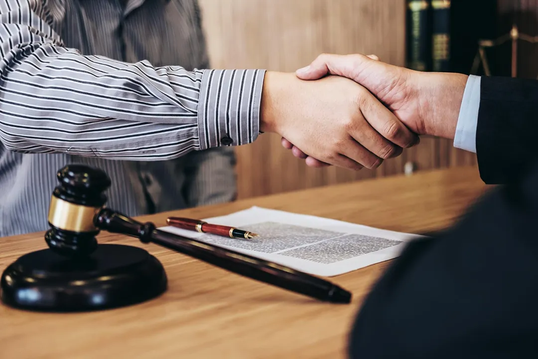 Two individuals shaking hands over a wooden desk with legal documents, a pen, and a judge's gavel.