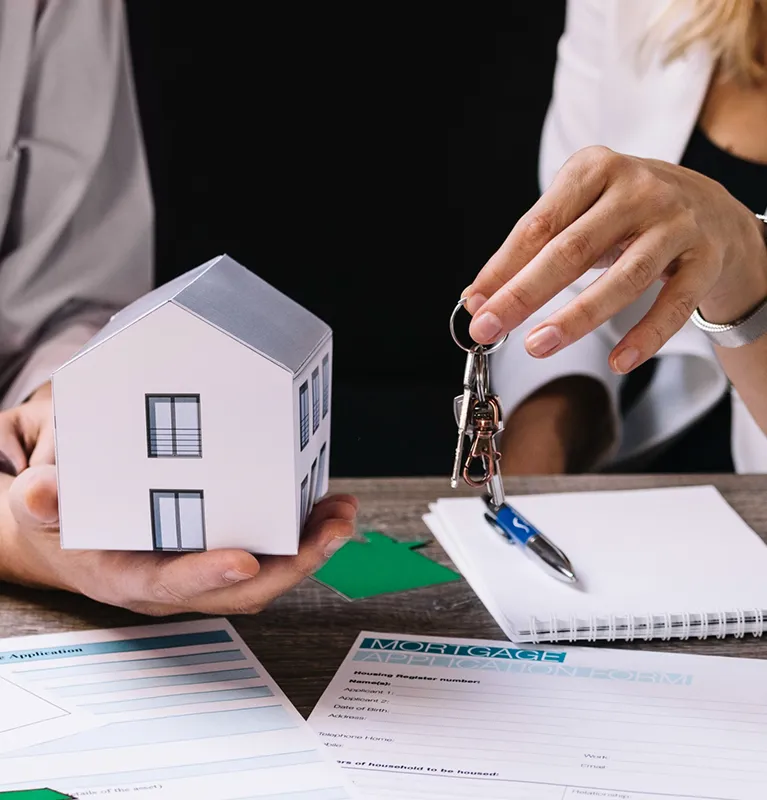Two people holding a small house model and keys over mortgage application documents.