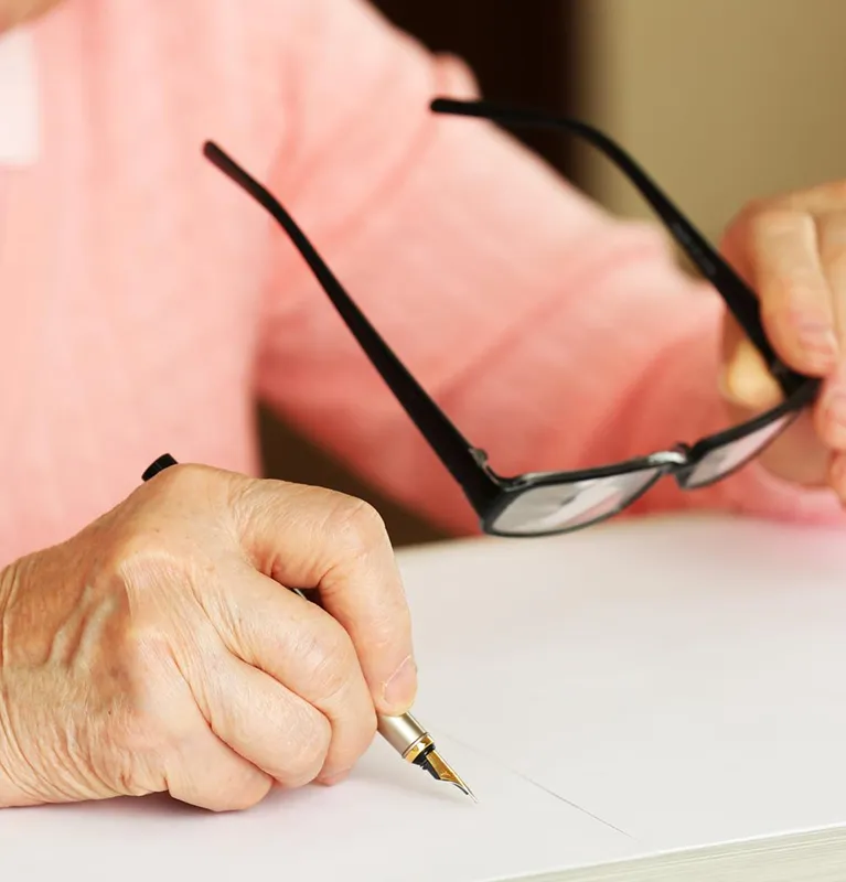 Close-up of elderly hands holding a fountain pen and black eyeglasses over white paper.