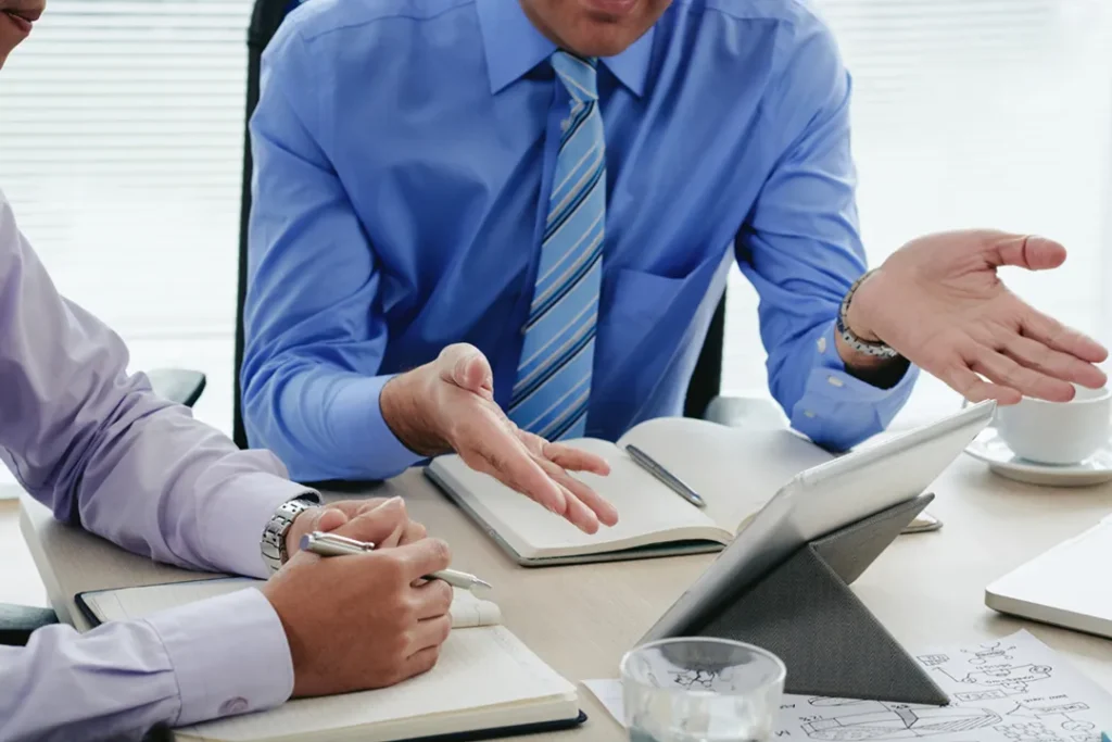 Two professionals in dress shirts and ties discussing while referencing a tablet and taking notes.