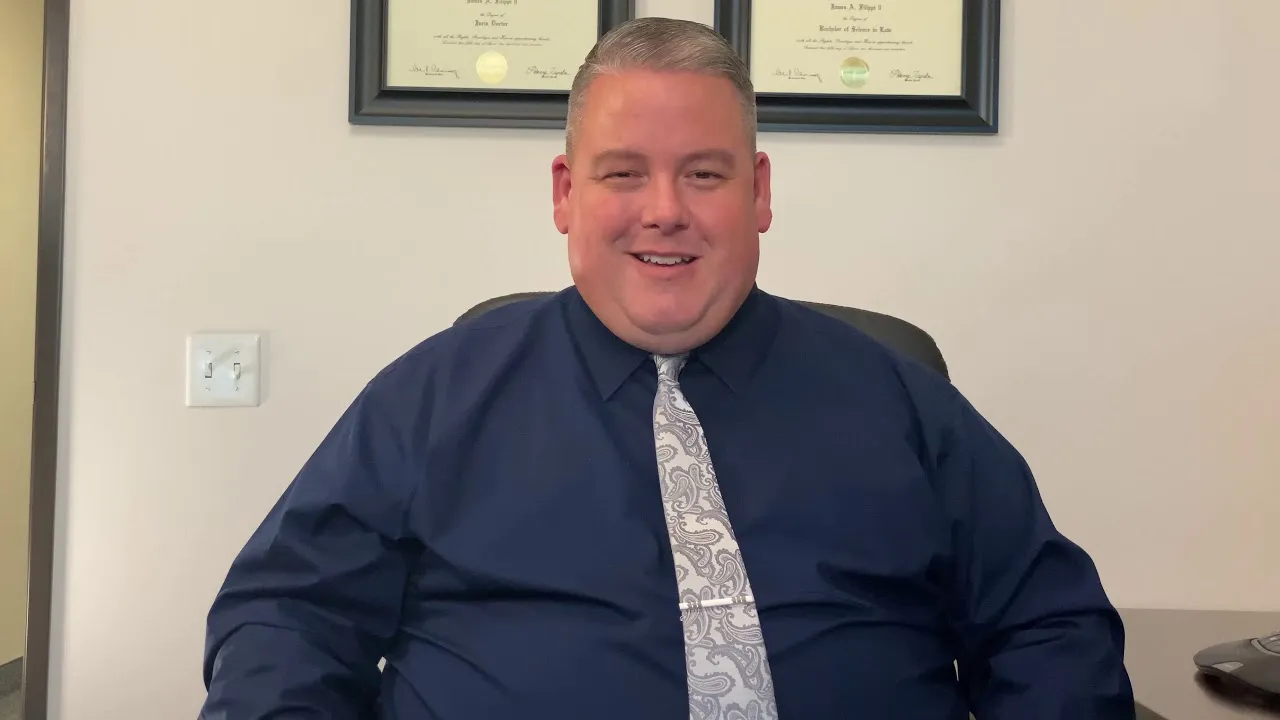 Middle-aged man in navy shirt and patterned tie seated in office with framed diplomas on wall.