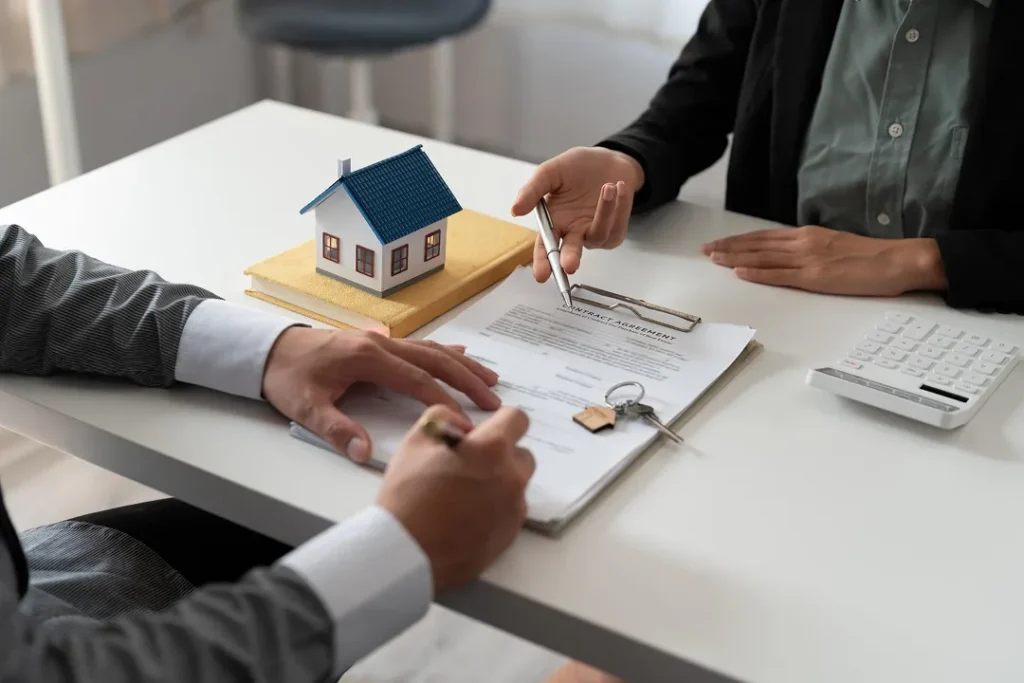 Two individuals at a desk reviewing a contract agreement with house keys and a model house.