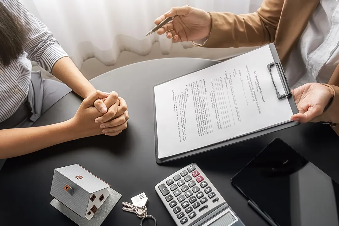 Two people discussing a contract over a black table with a house model, keys, calculator, and tablet.