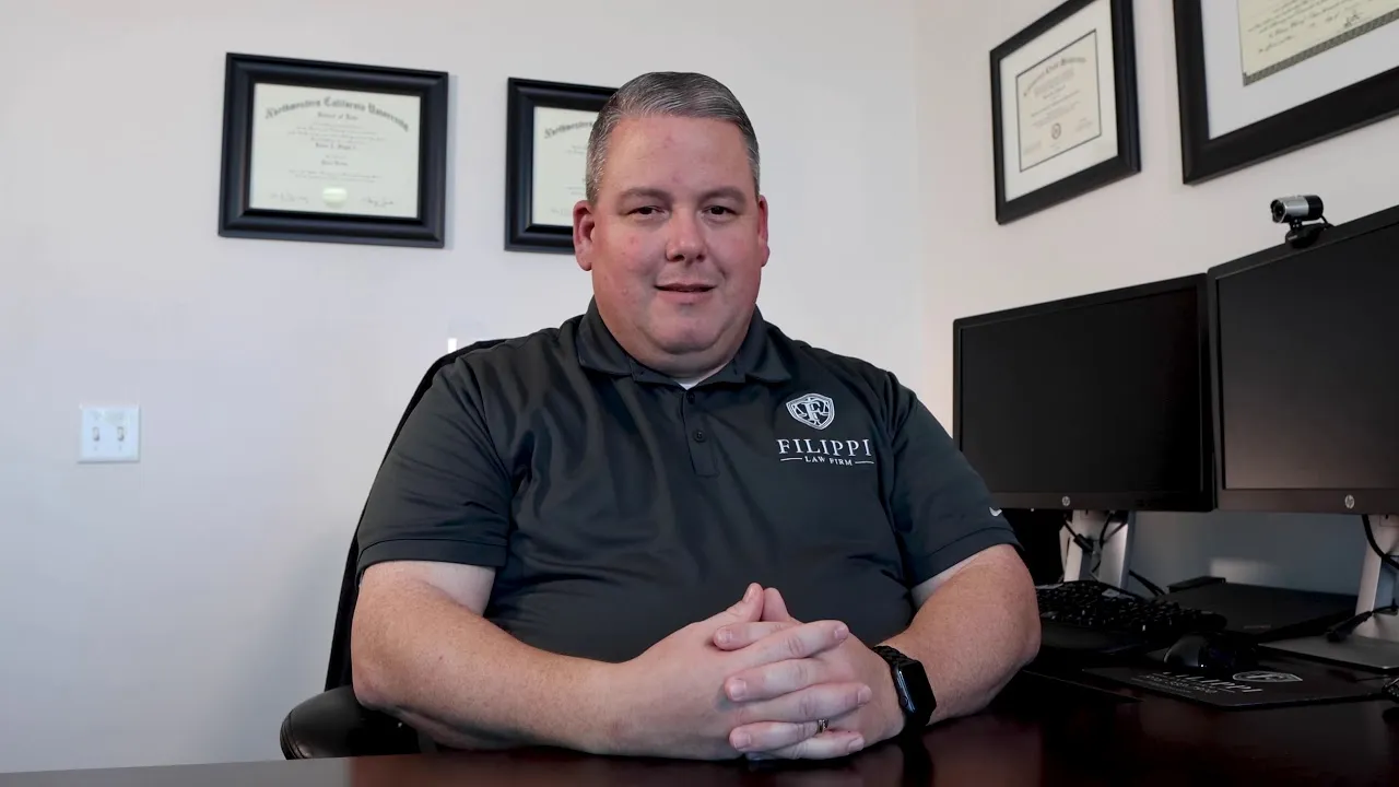 Professional man in black polo seated at desk with dual monitors and framed diplomas on wall.