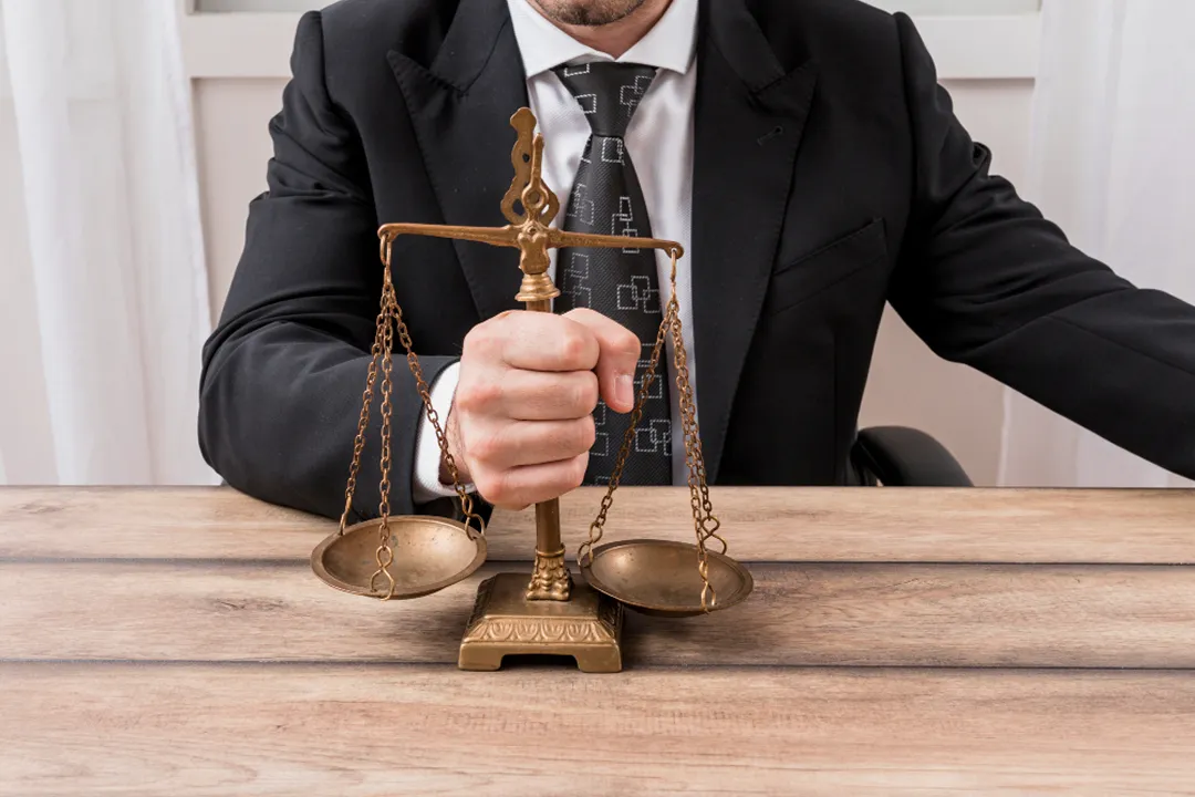 Man in black suit holding antique brass balance scales on wooden table.