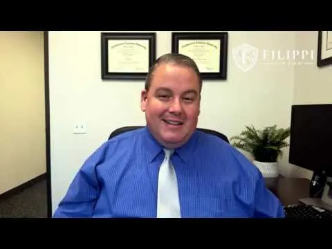 Middle-aged man in blue shirt and white tie sitting in office with framed certificates and plant.