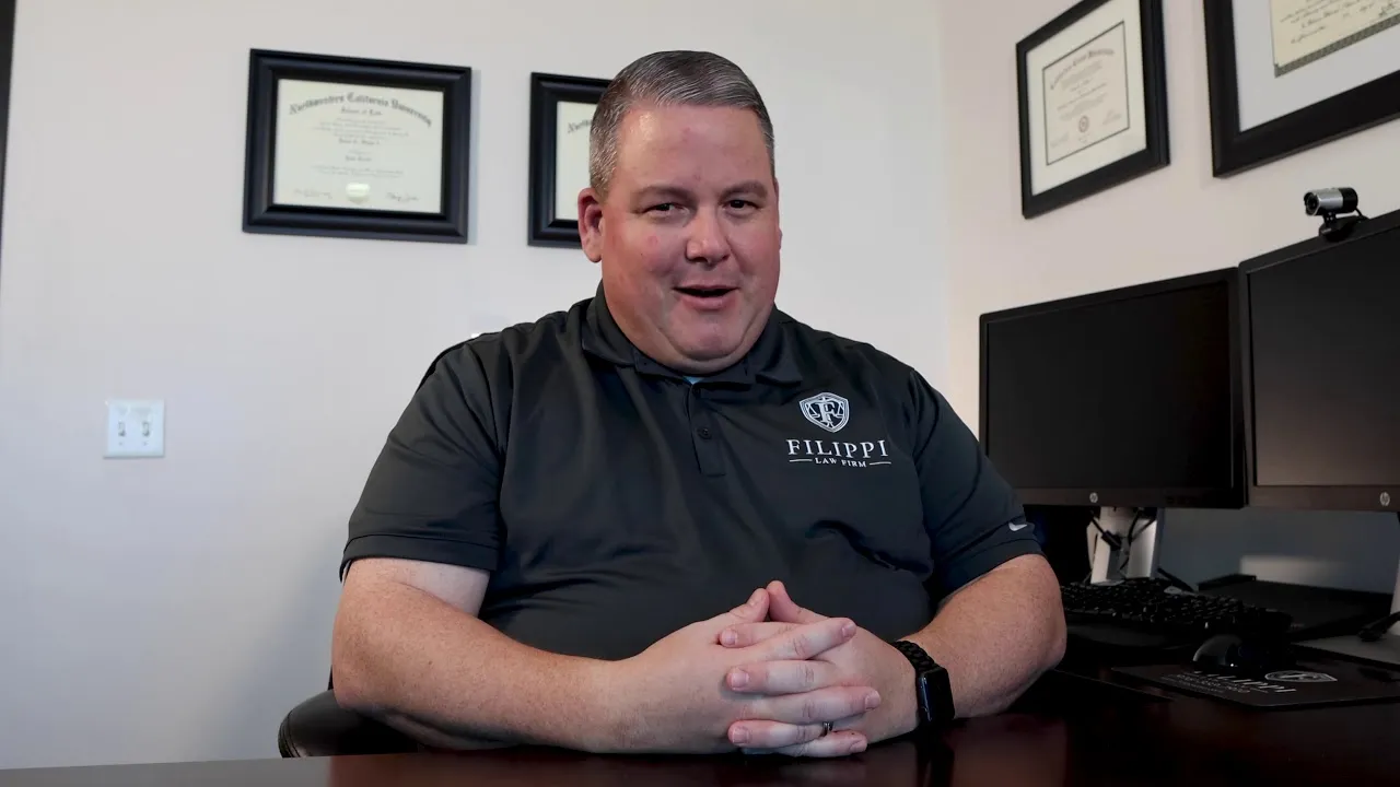 Middle-aged man in black polo shirt with law firm logo seated at desk, diplomas on wall.