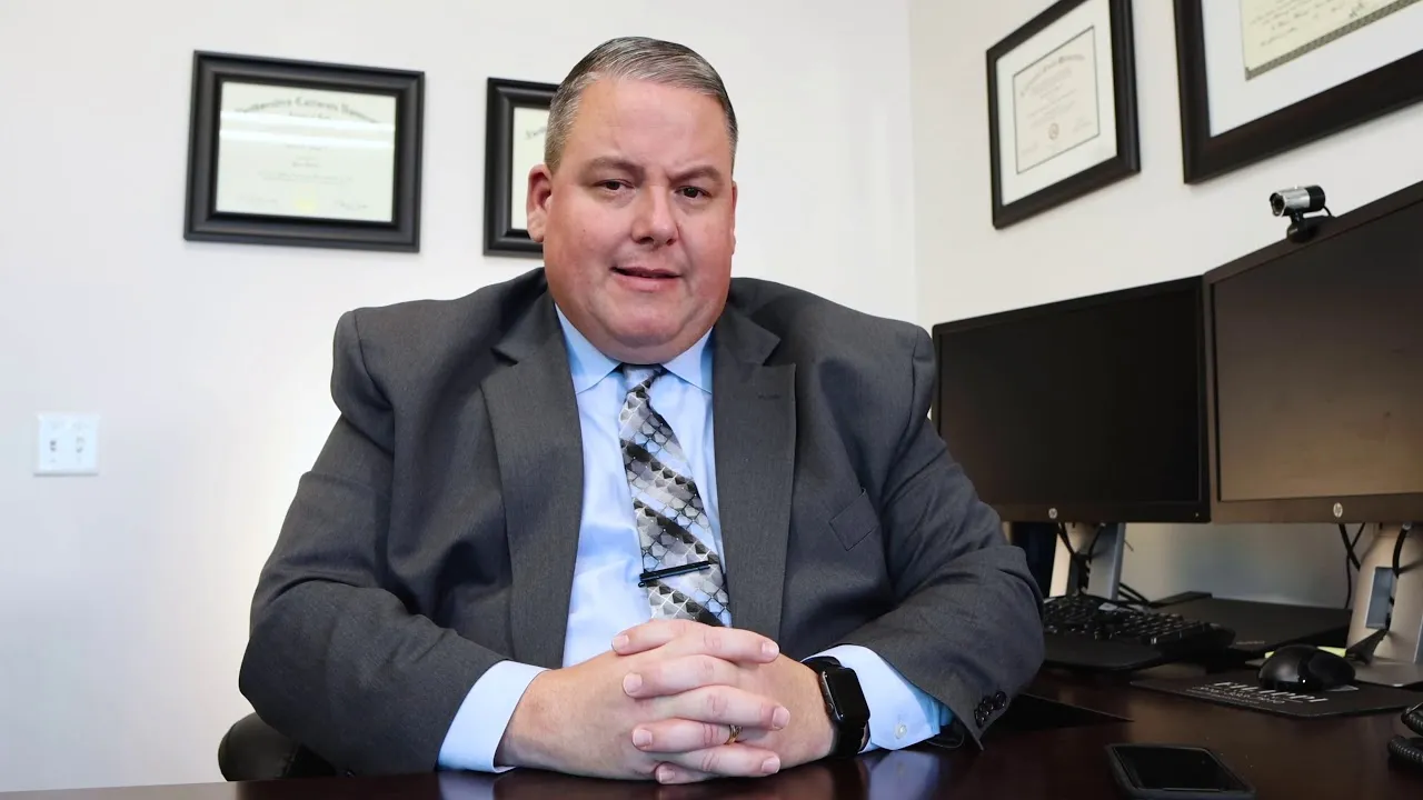 Professional man in gray suit seated at desk with dual monitors and framed certificates on wall.