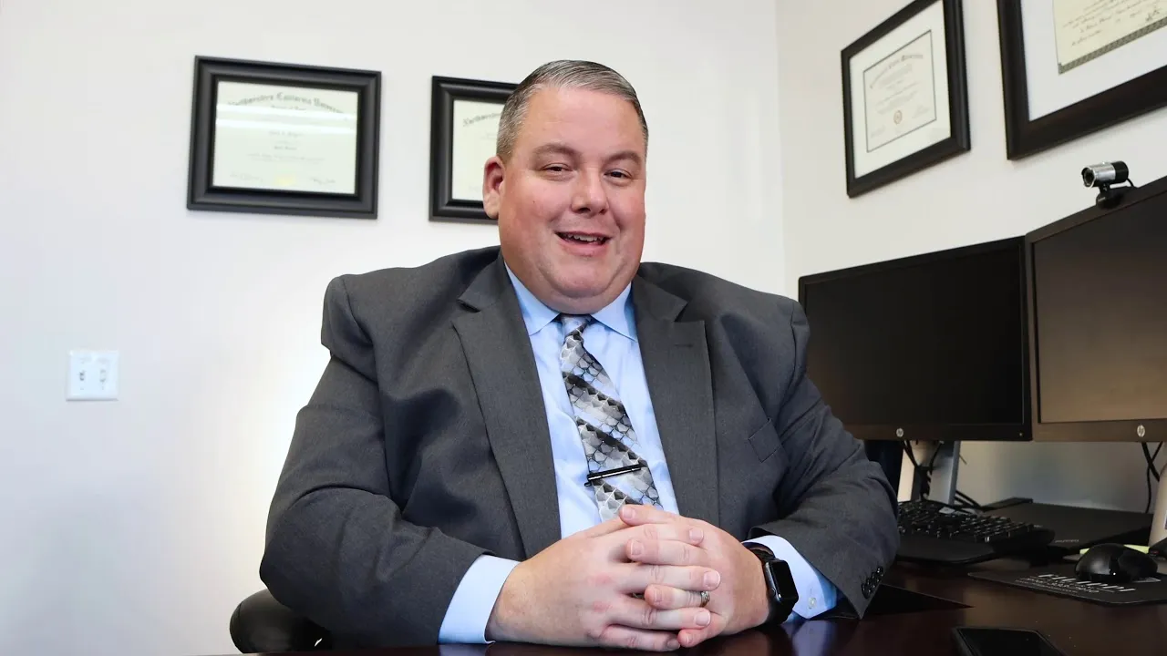 Professional man in gray suit seated at desk with dual monitors and framed certificates on wall.