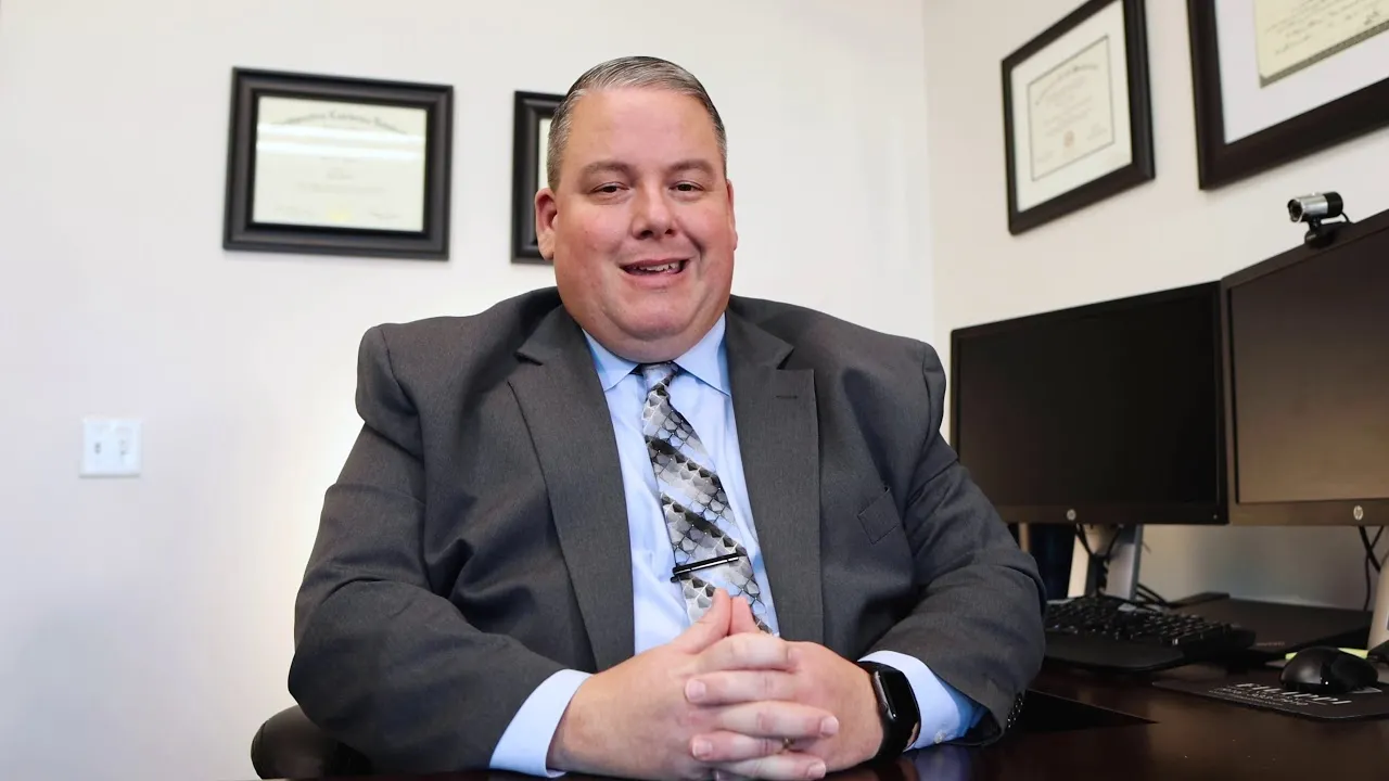 Professional man in gray suit and patterned tie seated at desk with computer monitors and framed certificates on wall.