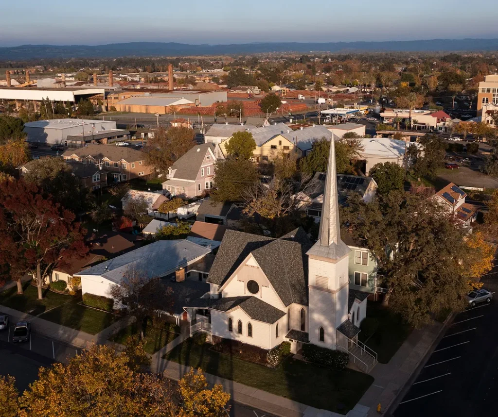 Aerial view of a white church with a tall steeple surrounded by residential and commercial buildings during late afternoon.