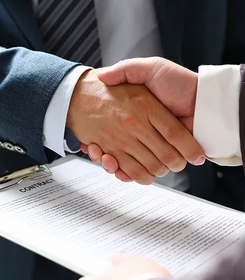 Two individuals in business attire shaking hands over a signed contract document.