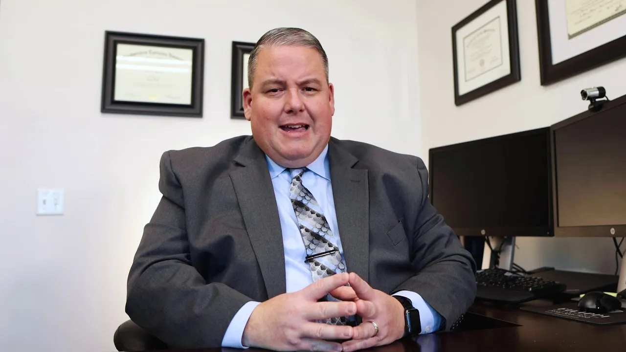 Middle-aged man in gray suit and patterned tie seated at desk with dual monitors and framed certificates on wall.