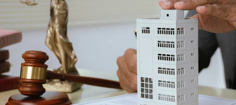 Hand placing a small white building model on a desk with a wooden gavel and legal books in the background.