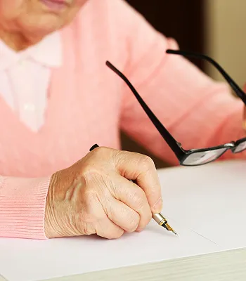 Elderly person in a pink sweater writing with a pen, holding black eyeglasses.