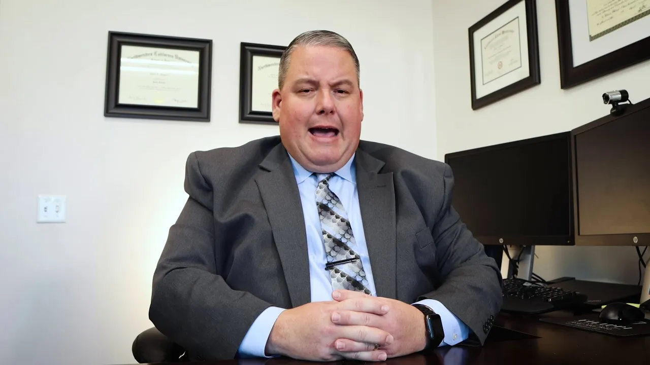 Professional man in gray suit and patterned tie seated at desk with dual monitors and framed certificates on wall.