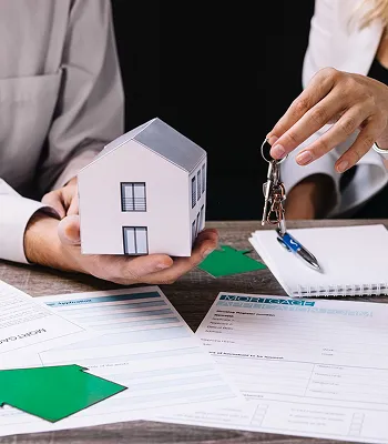 Two people at a table with a small white house model, keys, and real estate documents.
