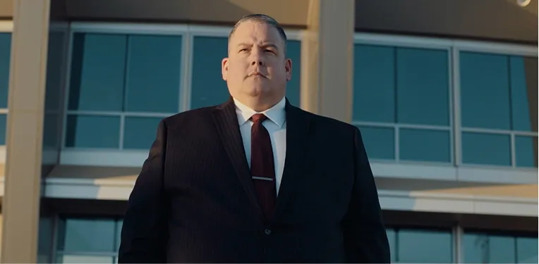 Middle-aged man in dark suit and burgundy tie standing confidently outdoors by modern glass building.