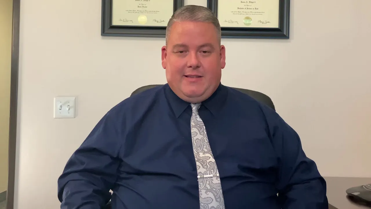 Middle-aged man in a navy shirt and paisley tie seated in office with framed diplomas on wall.