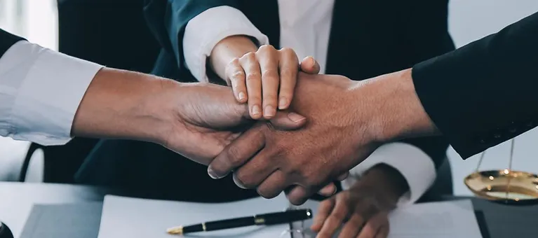 Three business professionals in formal attire placing hands together over a desk with documents and a pen.