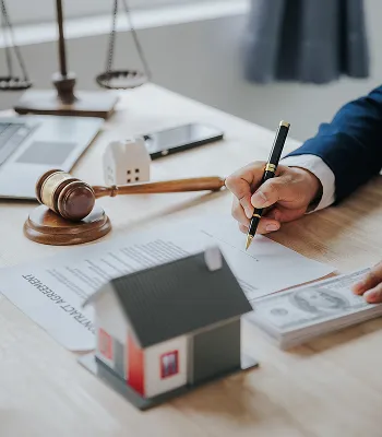 Person signing legal documents at desk with gavel, miniature house model, and money.