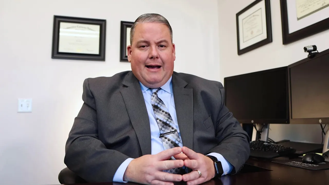 Middle-aged man in gray suit and patterned tie seated at desk with dual monitors and framed certificates on wall.