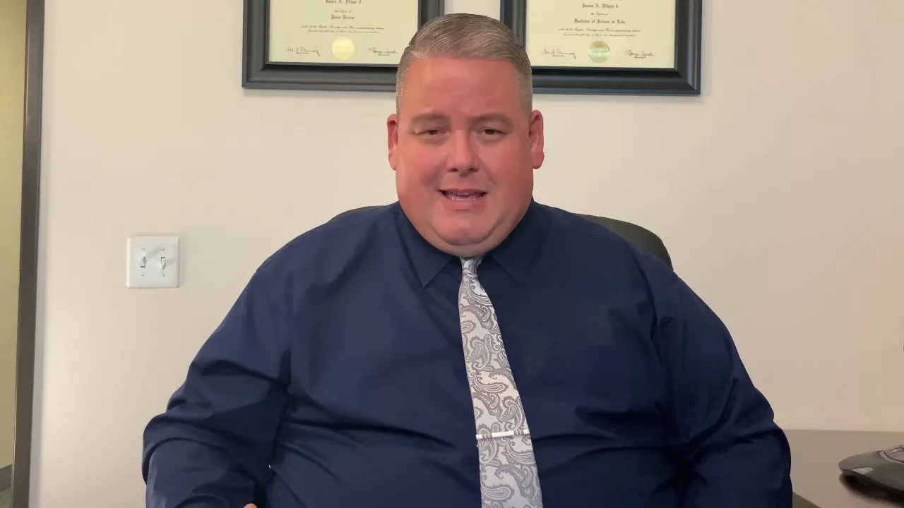 Middle-aged man in navy shirt and patterned tie seated in office with framed diplomas on wall.