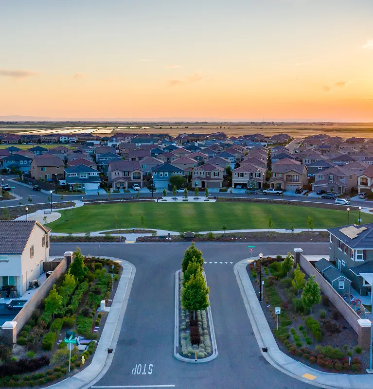 Aerial view of a suburban neighborhood at sunset with houses, green lawns, and streets.