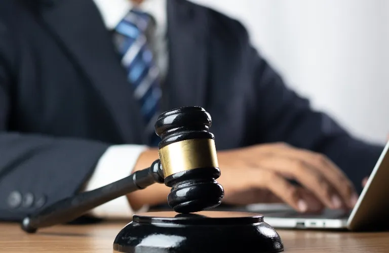 Close-up of a black judge's gavel with brass band on wooden table, person in suit typing on laptop in background.
