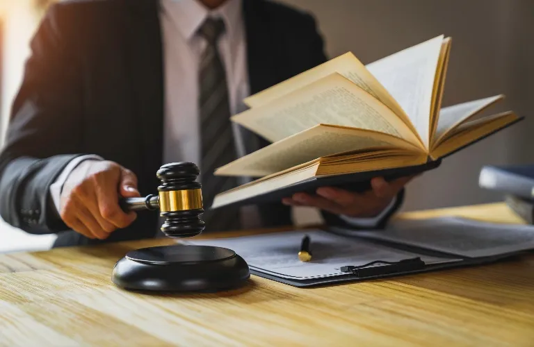 Person in business attire holding a gavel and an open book over a wooden desk.