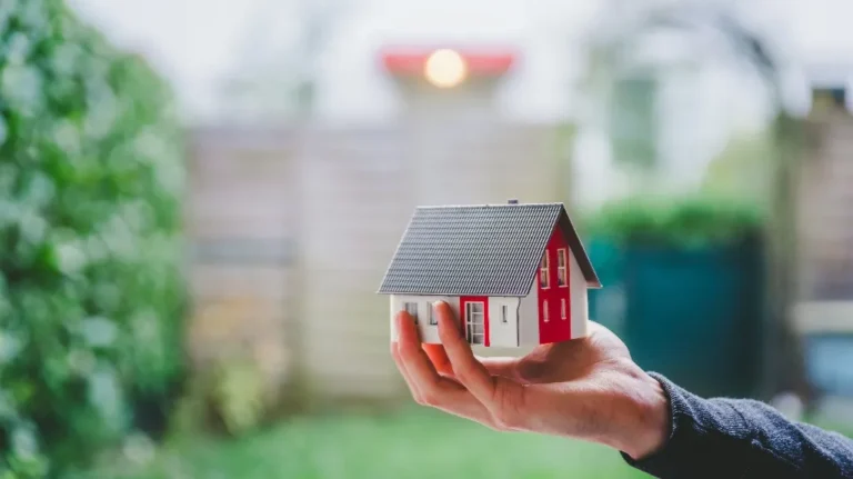 Hand holding a small model house with a gray roof and red and white walls outdoors.