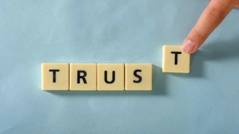 Hand placing a letter tile "T" to complete the word "TRUST" on a light blue surface.