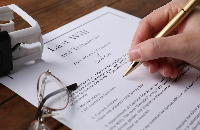 Close-up of a hand holding a gold pen, signing a last will and testament document on a wooden table.