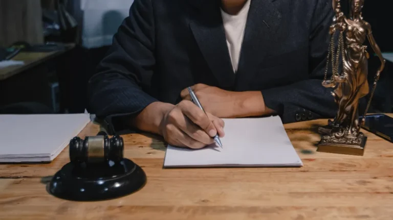 Person in dark suit writing on paper at wooden desk with gavel and Lady Justice statue.