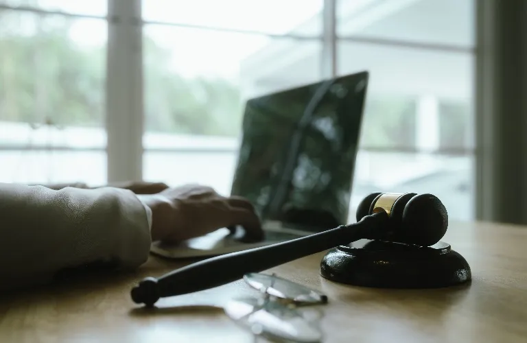 Wooden judge's gavel on desk with eyeglasses and person using laptop in background.
