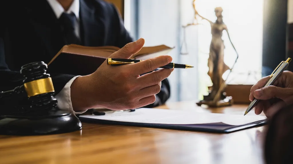 Two individuals in formal attire discussing legal documents with a judge's gavel and Lady Justice statue on a wooden desk.
