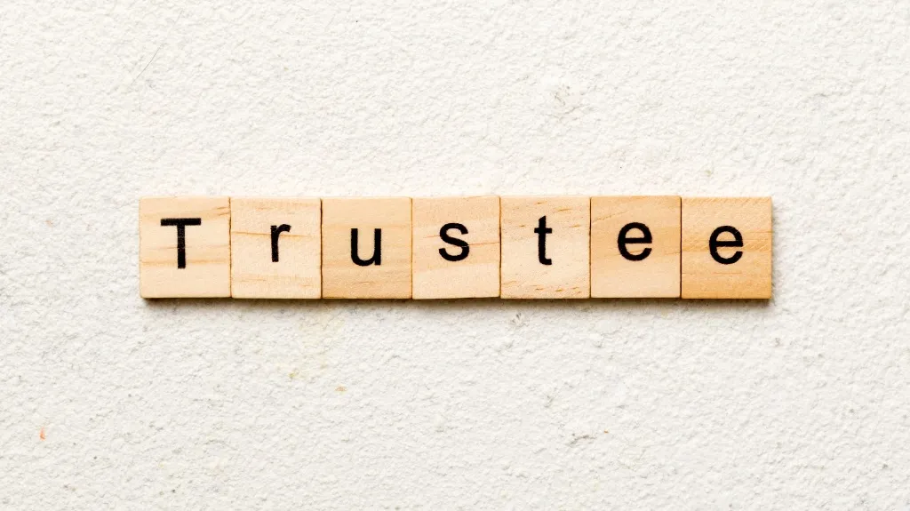 Wooden letter tiles arranged to spell the word "Trustee" on a textured white surface.